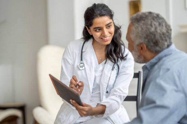 A young female doctor of Middle Eastern decent, sits with a senior patient in the comfort of his own home during a Homecare visit.  She is wearing a white lab coat, has a stethoscope around her neck and is holding out a tablet as she shows the gentleman his recent test results.   The senior is leaning in to look at the screen as he listens attentively.