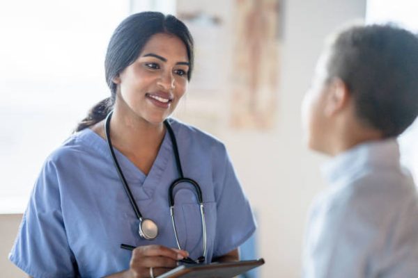 A young mixed race boy sits up on an exam table as he answers some questions the nurse has for him.  He is dressed casually and facing the nurse who is seated in front of him.  She is wearing blue scrubs and recording the young boys answers on a tablet she has in her hands.